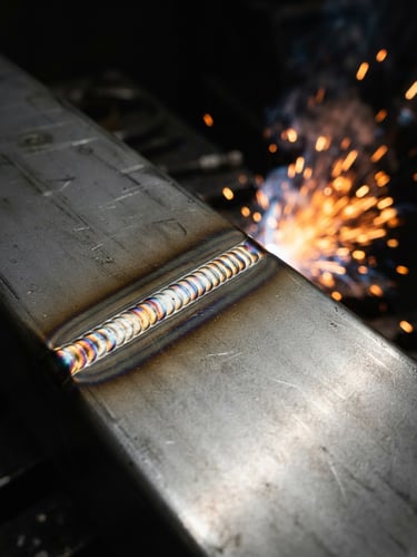 Macro shot of clean, high-tolerance TIG weld bead on stainless steel joint, high-contrast industrial shop lighting, glowing orange sparks in the background