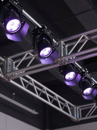 Detailed close-up of structural metal trusses and purple theater spotlights rigged to a dark ceiling, technical precision.