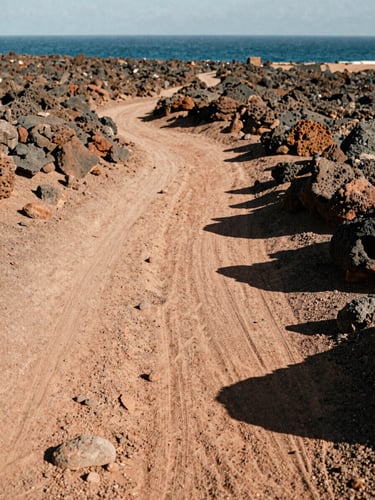 Warm editorial photography of a dusty desert path leading to the sea in Cabo de Gata, harsh midday sun casting deep shadows, volcanic rocks, 35mm film style.