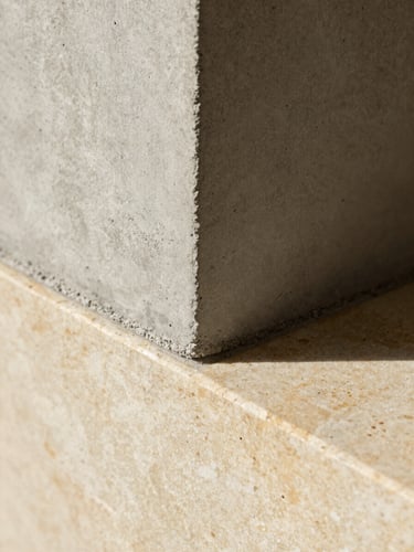 Extreme macro architectural photograph showing the precise material transition where raw board-formed concrete meets smooth, warm travertine stone, natural side light casting soft shadows.