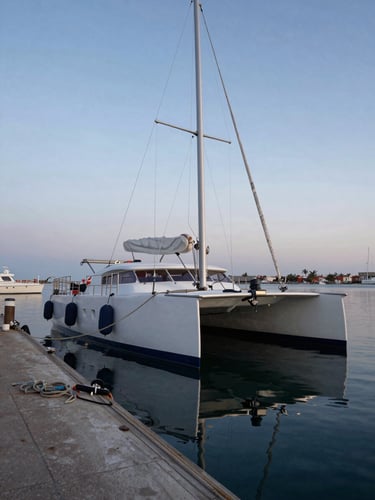 Environmental wide shot from the dock looking along the hull of a catamaran moored at a small marina in La Paz, morning blue hour light, ropes and fenders in the foreground, calm water reflecting the hull, no people visible, documentary framing
