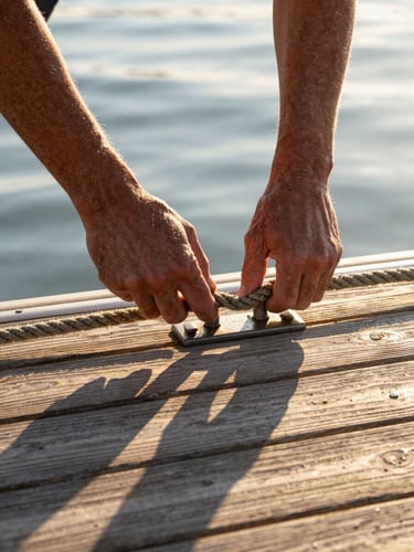Close portrait framing from water level — weathered hands securing a dock line to a cleat on a wooden deck, golden hour light casting long shadows across the teak grain, rope fibers visible in detail, no faces, just the hands and the hardware in motion