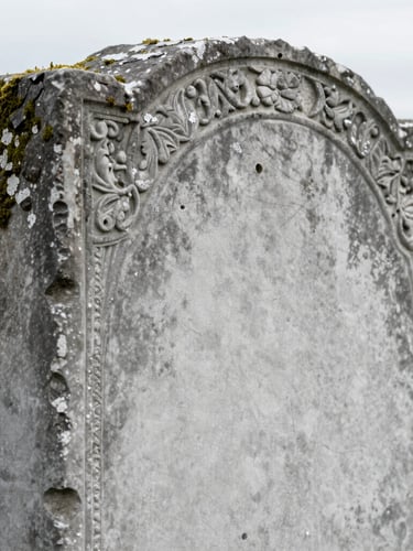 Documentary photograph of a historic gravestone, showing the clean boundary line between the weathered, dark moss-covered side and the restored, clean light grey stone side, soft overcast daylight.