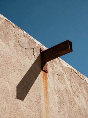 Low-angle architectural shot of a weathered stucco wall meeting a rusted steel beam, harsh midday desert sun casting a deep sharp shadow against a clear deep blue sky.
