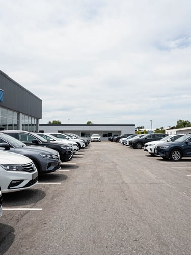 Wide environmental shot of an open car dealership lot in bright overcast daylight, rows of vehicles parked under a pale sky, empty asphalt forecourt stretching to the right, dealership building facade visible at left edge, no people