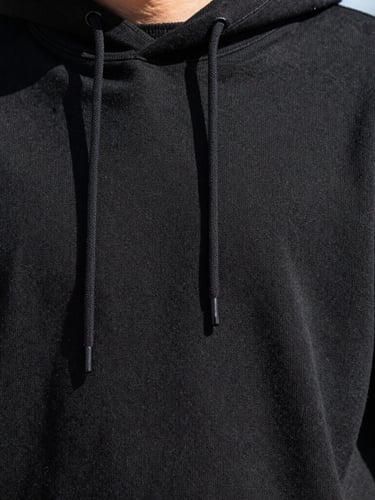 Close-up fabric texture detail of a high-quality heavy cotton black hoodie, showing clean stitching and premium weave, bright direct daylight, high-contrast, modern youth aesthetic