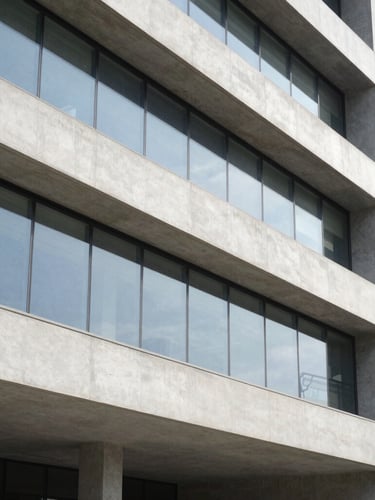 Detalhe arquitetônico de uma estrutura de concreto polido e vidro, com reflexos geométricos, sob luz natural nítida, enquadramento vertical.