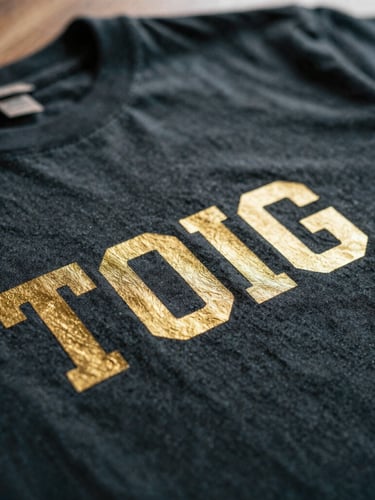 Macro close-up of custom metallic gold printing on a deep charcoal combed cotton t-shirt, showcasing the fine fabric texture and crisp print edges under warm window light, shallow depth of field, 35mm.
