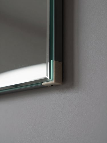 Extreme close-up of a custom mirror corner detail, frameless polished edge meeting a wall-mounted concealed bracket, soft studio light from upper left revealing the precise seam between glass and substrate, muted gray plaster background, no people, tight 45-degree angle showing the mount geometry and backing material transition