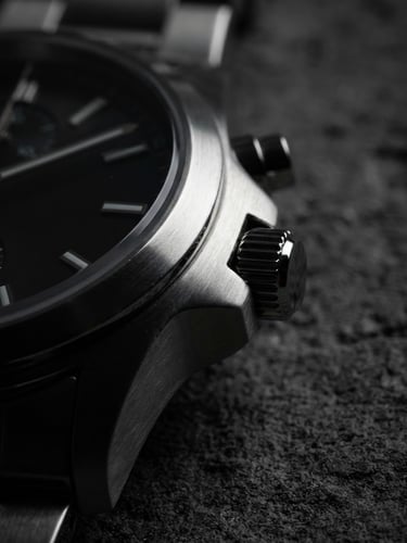 Sharp macro product photography of a brushed steel watch case catching directional light at three o'clock, deep shadows, architectural concrete background