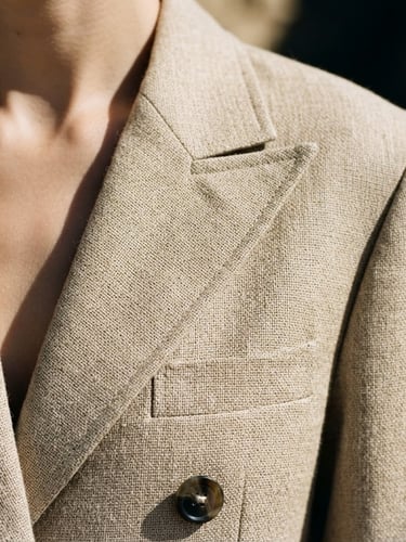 A close-up high-fashion crop of structured, sand-colored hemp tailoring. Sharp studio lighting casts dramatic shadows across the clean, architectural lines of the lapel.