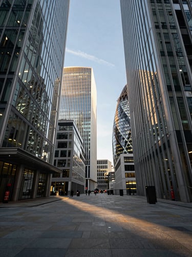 Wide environmental shot from street level looking up at glass-and-steel towers in London's financial district, late afternoon daylight casting long shadows across a polished stone pavement, no people in the immediate foreground, cool sky with faint golden light catching upper floors