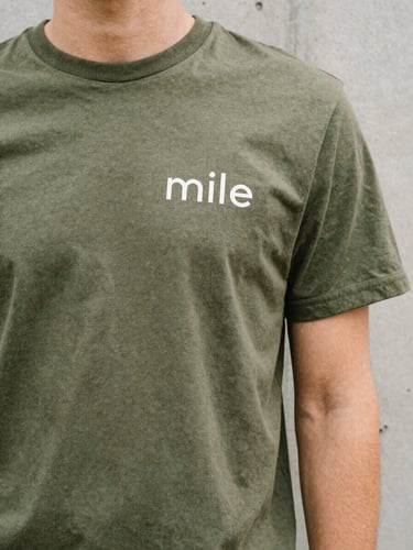 A candid close-up of a person wearing a premium olive green t-shirt with clean minimalist typography, standing in soft overcast daylight next to a concrete wall.
