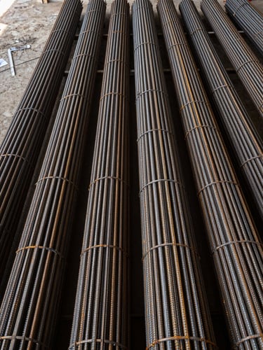 Overhead wide shot looking straight down at tightly packed rebar bundles arranged in a warehouse supply yard, steel surfaces showing mill scale and galvanized sheen, diffused loading-dock daylight from the left edge, no human presence, framing emphasizes repetition and stock depth
