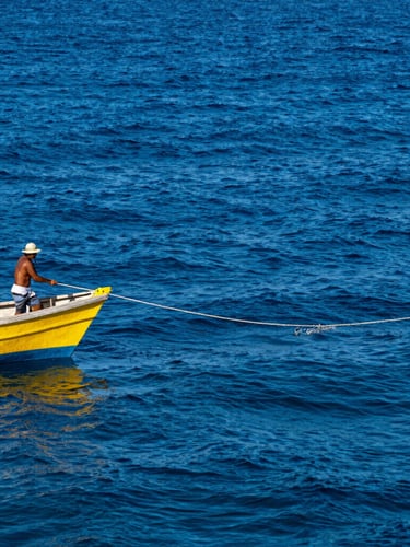 A local fisherman on a small wooden vessel pulling a thick handline from deep blue water, harsh tropical sunlight, sharp focus