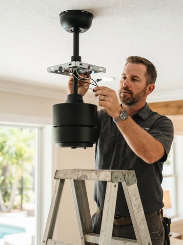 A close-up of a professional handyman installing a modern black ceiling fan in a bright Florida living room, warm daylight, shallow depth of field