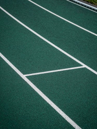Close-up overhead view of a running track's lane markings on rubberized surface, crisp geometric lines in deep forest green and white against charcoal base, studio-controlled cool light emphasizing material texture and spatial precision, no human presence