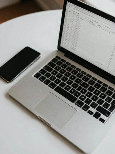 Overhead close-up of an open laptop on a clean desk showing a spreadsheet, a smartphone beside it, cool north-facing daylight, minimal Colombian home-office environment