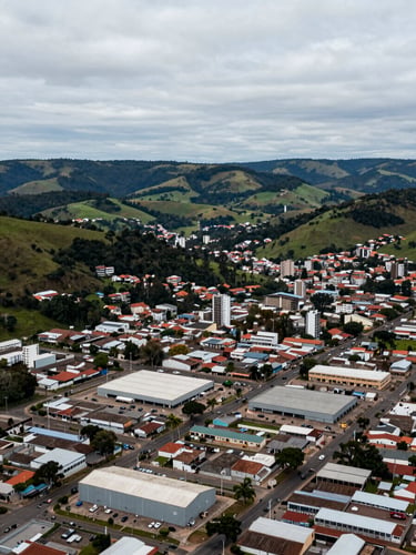 Wide panoramic aerial view of a small southern Brazilian city surrounded by Serra Gaúcha hills and green valleys, industrial and commercial district visible in the foreground with warehouse rooftops, overcast cool daylight giving a neutral industrial tone, horizon line low, generous sky above, no text or signs legible