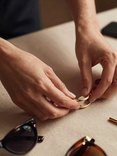 Close-up of hands meticulously arranging fashion accessories on a beige fabric, soft studio lighting, rich muted tones, elegant styling, shallow depth of field, warm editorial fashion photography.
