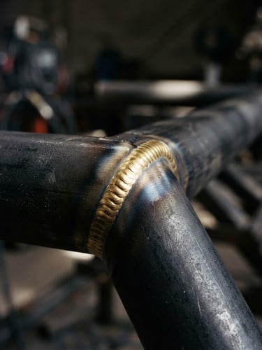 A high-contrast close-up of a flawless brass weld joint on a deep charcoal steel frame, sharp focus, industrial editorial style, 35mm lens.
