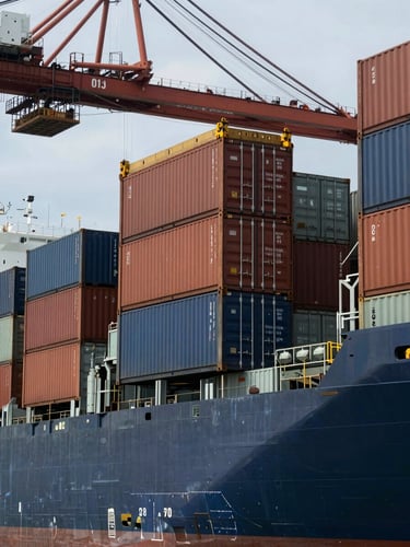 Close-up wide panoramic view of shipping containers being loaded onto a cargo vessel at an Indonesian port, container crane visible upper left, overcast cool daylight, no people in foreground, industrial scale and organized logistics visible, dark steel and brown container surfaces