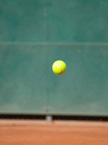 Dynamic close-up action shot of a tennis racket hitting a yellow tennis ball mid-air, red clay dust exploding from the impact, bright sunny afternoon lighting, shallow depth of field.