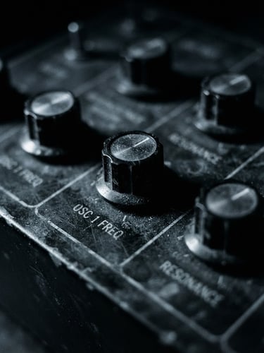Extreme close-up of vintage synthesizer knobs in dim, high-contrast chiaroscuro lighting, desaturated monochrome with cold blue undertones, grainy 35mm film.
