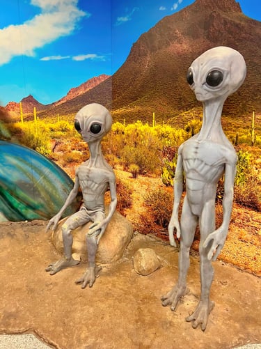 Two grey alien statues at a Roswell UFO exhibit with a desert mountain landscape background.
