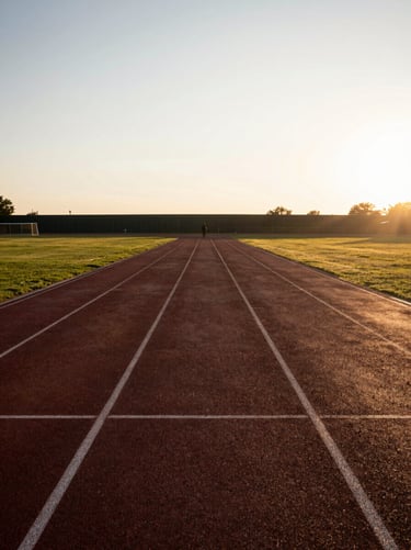 Wide environmental shot of an empty athletics track at early-morning golden hour, a lone figure standing still at the far end in silhouette against low raking light, long shadows stretching toward camera, contemplative and precise atmosphere, no action or motion blur
