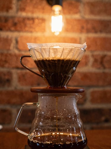 Close-up of a glass pour-over coffee dripper with freshly brewed coffee dripping, warm amber backlight, exposed brick wall in soft focus
