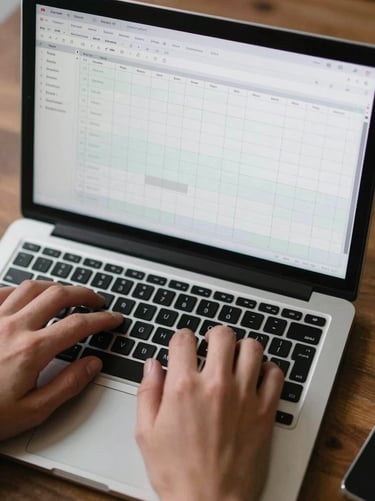 Close-up overhead shot of hands on a laptop keyboard, a spreadsheet with weekly milestone rows visible on the screen, screen glow as the primary light source, desk surface visible at the edges, no faces, documentary framing