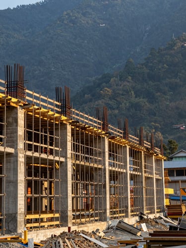 Wide environmental shot of a construction site at golden hour, scaffolding rising against a deep blue-green mountain backdrop in Nepal, long horizontal sightline showing scale of the project, natural warm light raking across concrete and timber framing