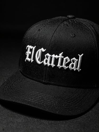 High-contrast black and white close-up of a black structured snapback cap with white gothic embroidery of El Cartel on the front, sharp studio lighting.