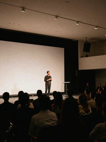Wide environmental shot of a modern keynote stage in Los Angeles, soft stage lighting illuminating a speaker with an attentive audience in silhouette