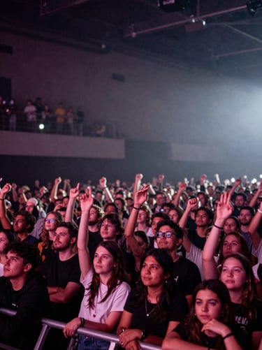 A wide-angle shot of a cheering crowd in a dimly lit venue, seen from the stage perspective. Hints of deep mauve and misty white lights illuminate the fans. Dramatic and engaging concert atmosphere.
