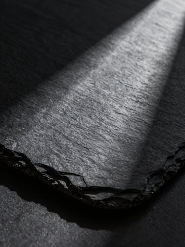 Extreme close-up of a monolithic honed slate slab, sharp directional light cutting across the dark matte texture, deep chiaroscuro shadows, high contrast.