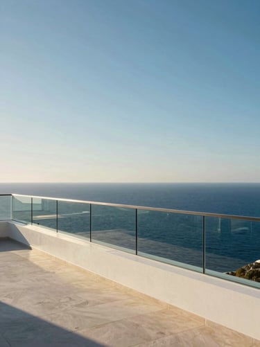 Wide panoramic view from a contemporary white Mediterranean terrace, frameless glass railing running along the left edge, the deep blue sea filling the horizon, golden midday sunlight flooding the stone floor, no furniture, vast empty sky above, minimal and serene