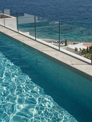Overhead study of a pool perimeter glass railing on a Costa del Sol rooftop, crystal-clear water below the glass panel, the Mediterranean sea visible beyond, midday light, no people, architectural and minimal