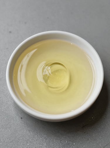 Macro overhead studio shot of pale-yellow BS Grade castor oil pooled in a small shallow dish, surface texture and viscosity visible, placed on a matte grey industrial surface, diffused overhead lighting, no shadows
