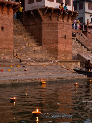 Ganges river bank in Varanasi at dusk, soft oil lamps floating on water, warm terracotta tones, documentary style, 35mm lens