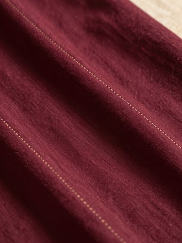 Close-up of premium wine-colored Kurti fabric showing high-thread-count cotton weave and delicate gold stitching details, warm editorial light