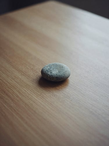 A single smooth stone on a wooden table, soft shadows, minimalist film photography, muted earthy tones, quiet contemplative mood, 35mm