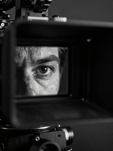 Monochrome close-up of a cinematographer's eye behind a camera viewfinder on a dark set, strong raking light from the right casting deep texture, high contrast, film grain, atmospheric shadow