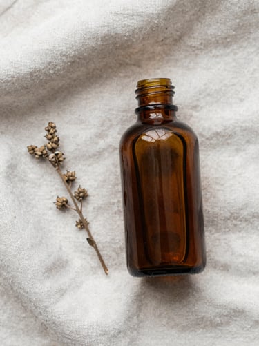 Close-up overhead flat-lay of a single amber glass apothecary bottle resting on a pale linen cloth, soft natural daylight from the left, one dried sprig beside it, no clutter