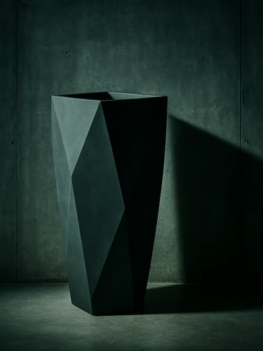Close-up of a tall, matte-charcoal geometric fiber planter standing against a raw concrete wall, casting a sharp shadow