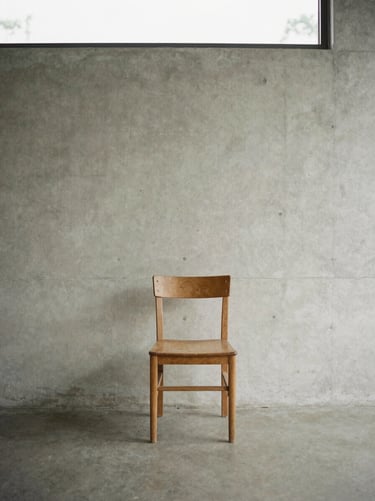 Minimalist editorial photography of a quiet interior with concrete walls and a single wooden chair, soft diffused overcast daylight, muted natural tones, medium format film, 80mm.