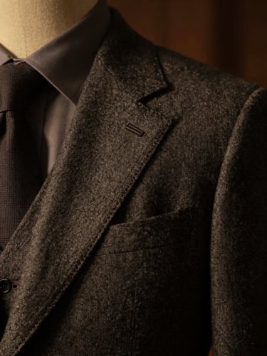 Close-up of a perfectly tailored charcoal wool suit jacket collar and hand-stitched lapel, dramatic side lighting, warm amber tones, shallow depth of field, dark moody photography, 35mm