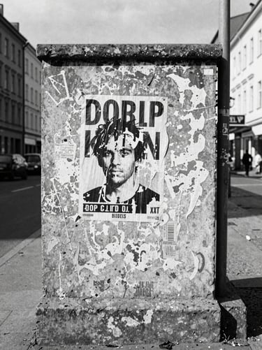 Gritty documentary film photography, harsh natural daylight, high-contrast monochrome, a weathered concrete wall with peeling posters in a shifting Berlin neighborhood, 35mm grain.