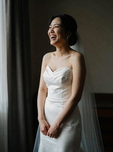 A bride laughing genuinely during preparation, natural window light, deep shadows, rich editorial photography.